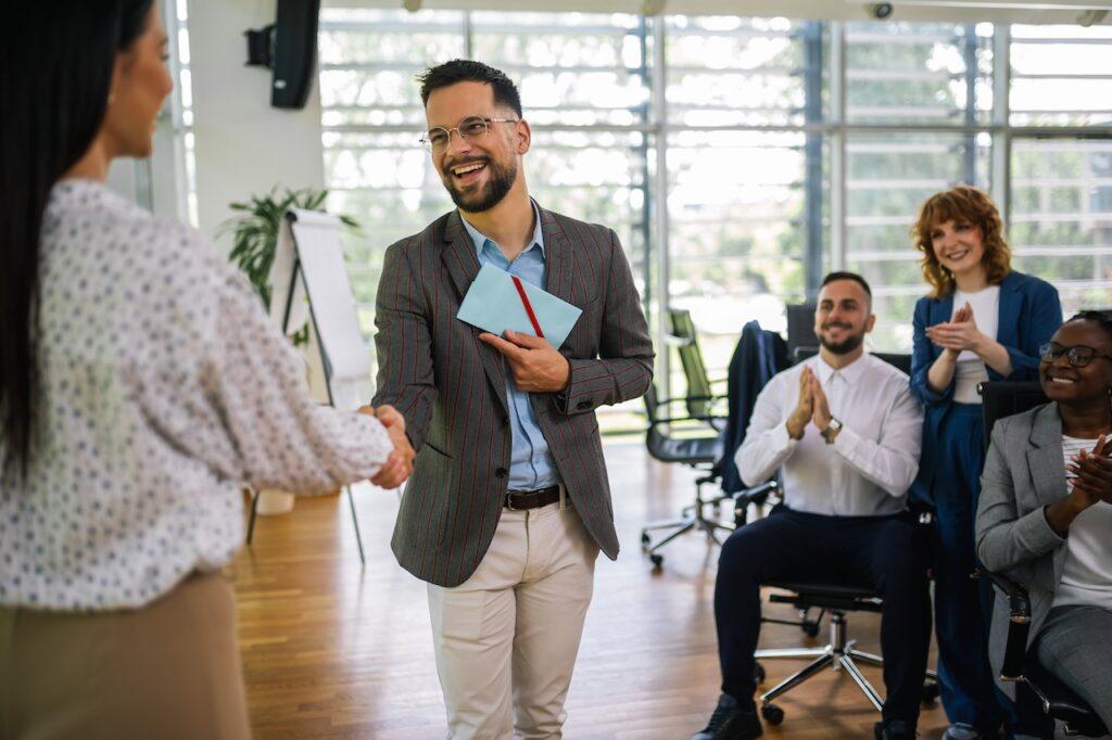 Ein Geschäftsmann überreicht einer Mitarbeiterin einen Umschlag mit einem Lächeln, während das Team applaudiert – Symbol für Wertschätzung und Belohnung im Arbeitsumfeld.