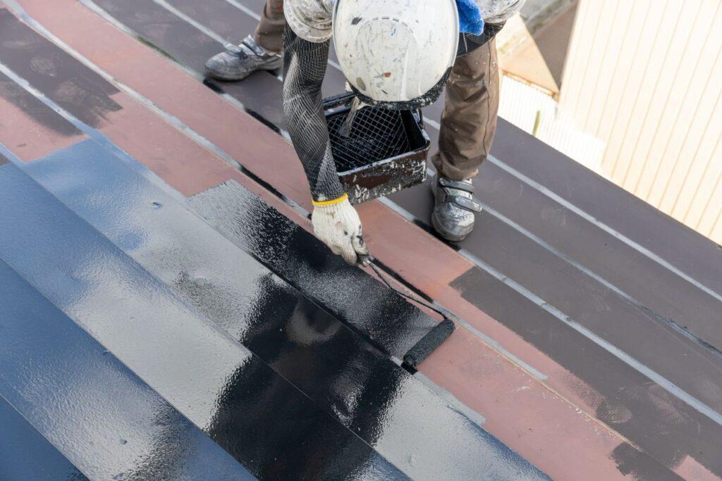 Handwerker traegt schwarze Schutzschicht auf Metalldach auf – professionelle Dachbeschichtung im Einsatz